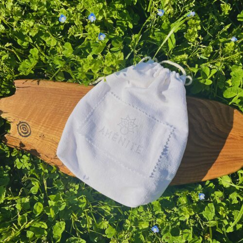 Petit pochon de transport en tissu blanc avec cordon de serrage, marqué du logo Aménité, posé sur une planche en bois dans l'herbe.