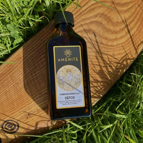 Flacon plat en verre ambré de la Synergie Hydrolats "Détox" Aménité, posé sur une planche en bois au milieu d'une herbe verte et dense sous un soleil vif.