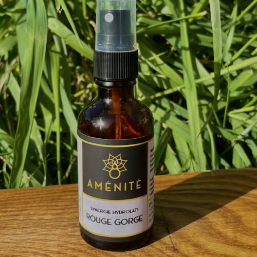 Flacon spray en verre ambré de la synergie d'hydrolats "Rouge Gorge" Aménité, posé sur un support en bois devant un fond d'herbes hautes éclairées par le soleil.