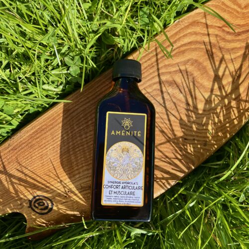 Flacon plat en verre ambré de la synergie "Confort Articulaire et Musculaire" Aménité, posé sur une planche en bois au milieu d'une herbe verte avec des ombres de feuilles ensoleillées