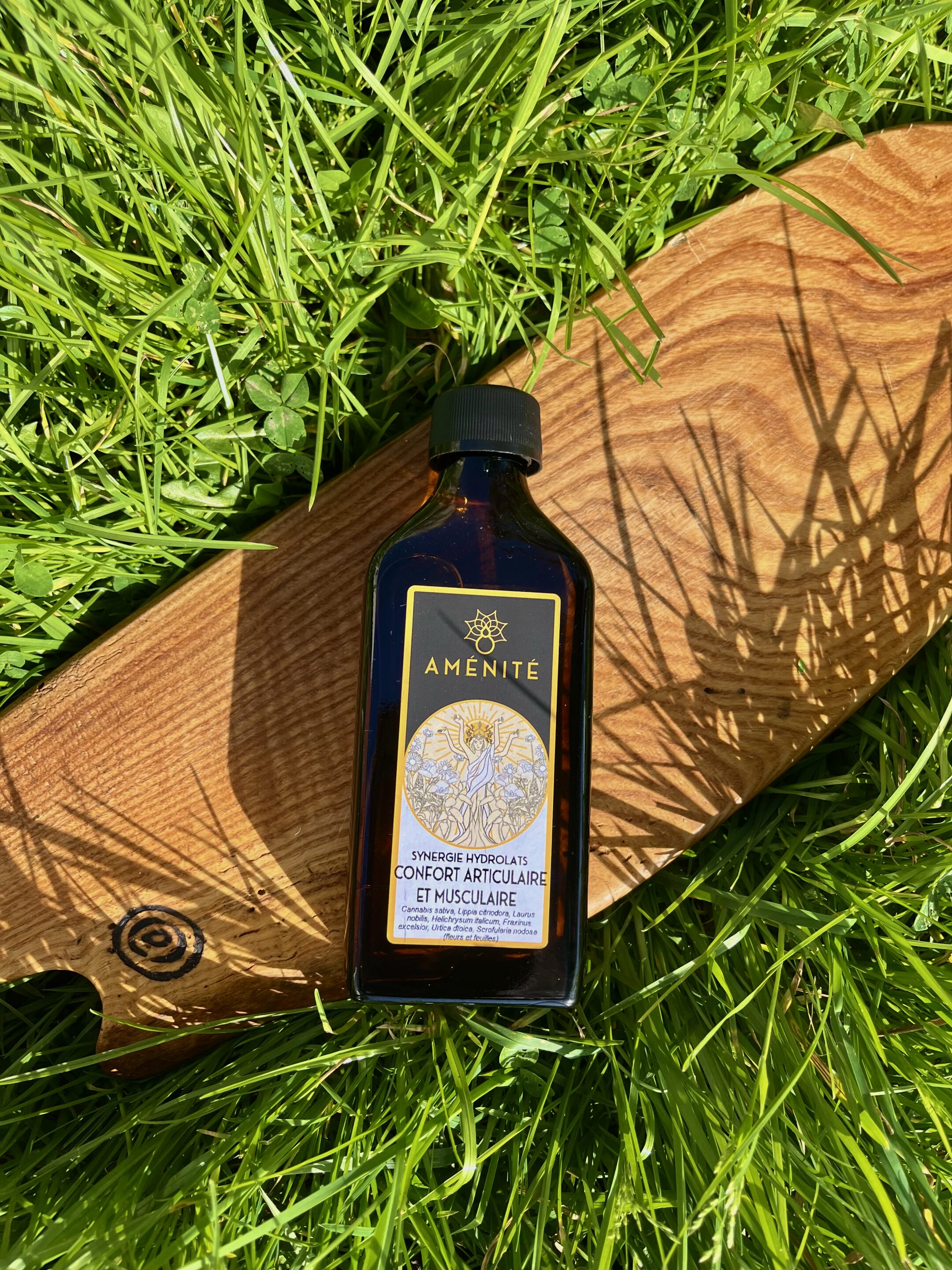 Flacon plat en verre ambré de la synergie "Confort Articulaire et Musculaire" Aménité, posé sur une planche en bois au milieu d'une herbe verte avec des ombres de feuilles ensoleillées