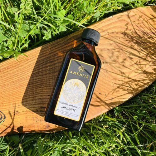 Flacon plat en verre ambré de la synergie d'hydrolats "Immunité" Aménité, posé sur une planche en bois clair au milieu d'une herbe verte et ensoleillée.