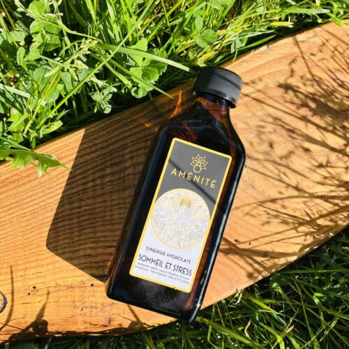 Flacon plat en verre ambré de la synergie "Sommeil et Stress" Aménité, présenté sur un support en bois dans un jardin ensoleillé avec des ombres de feuilles.