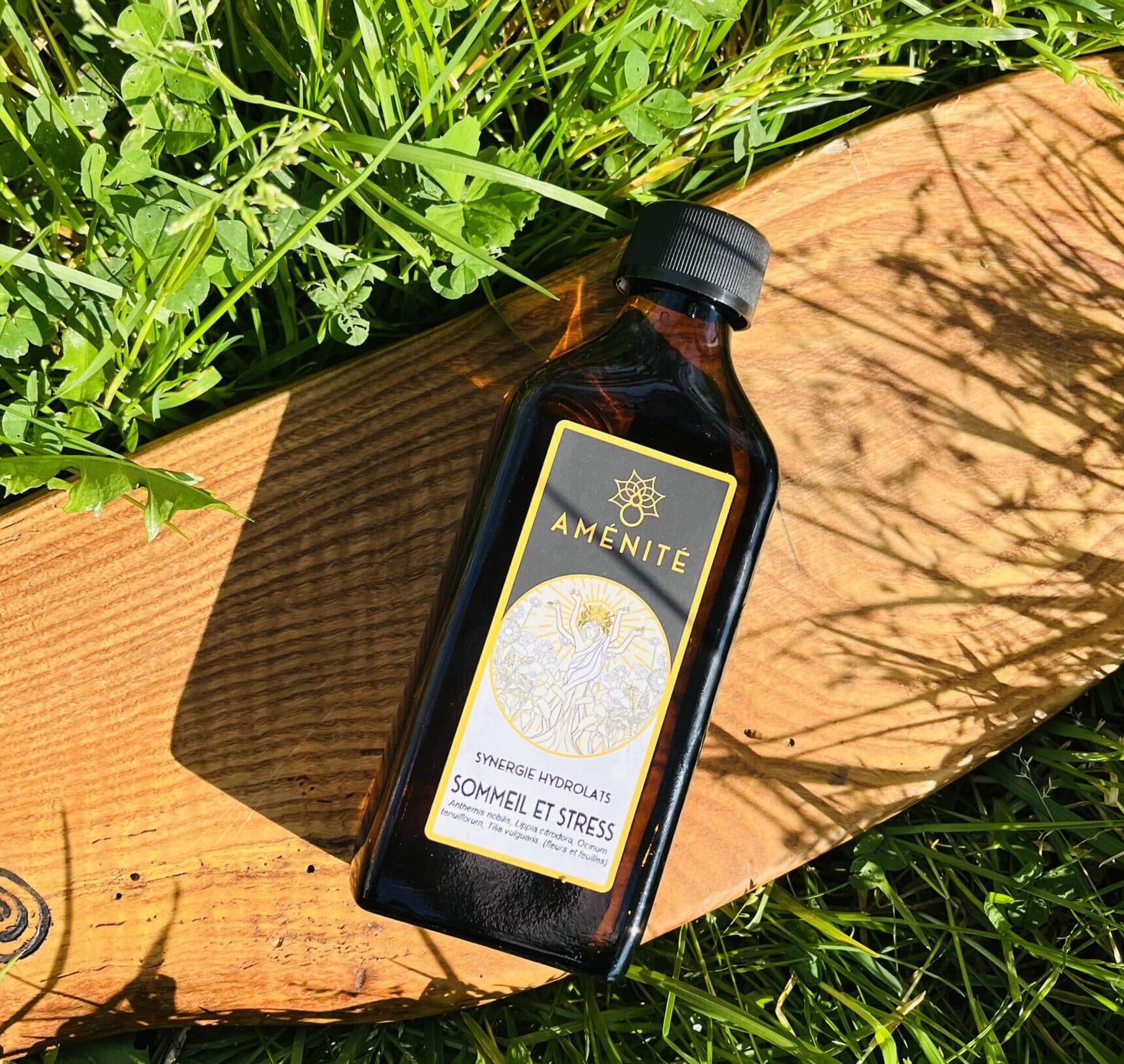 Flacon plat en verre ambré de la synergie "Sommeil et Stress" Aménité, présenté sur un support en bois dans un jardin ensoleillé avec des ombres de feuilles.