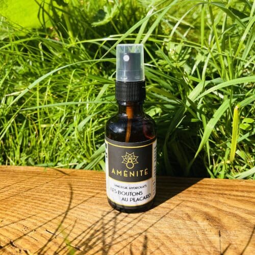 Flacon spray en verre ambré de la synergie "Les Boutons au Placard" Aménité, posé sur une planche en bois devant un arrière-plan d'herbes hautes vertes et ensoleillées.