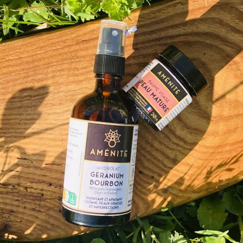 Un flacon spray d'hydrolat de Géranium Bourbon et un petit pot de Baume Visage Peau Mature posés sur un pochon blanc en tissu recyclé dans un jardin.