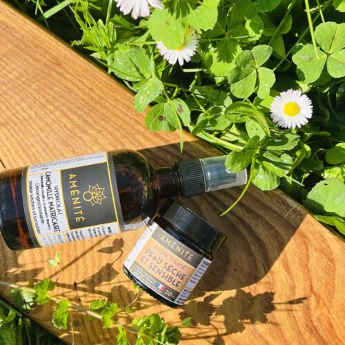 Hydrolat de Camomille Matricaire et Baume Peau Sèche et Sensible sur une planche en bois à côté de pâquerettes blanches.