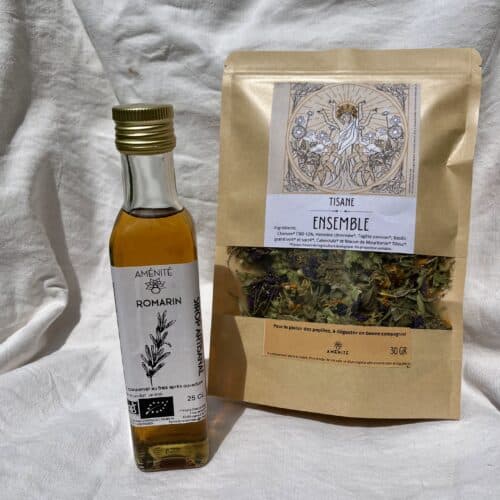 Sur un fond en tissu blanc froissé, une bouteille en verre de sirop artisanal est posée à côté d’un sachet kraft de tisane “Ensemble” contenant un mélange de plantes et fleurs séchées visibles à travers une fenêtre transparente.