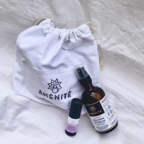 Pochette blanche brodée AMÉNITÉ contenant un hydrolat de géranium bourbon et un baume à lèvres naturel, posés sur un tissu blanc lumineux à l’ambiance douce et minimaliste.